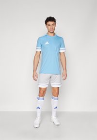 Svetlomodrý futbalový dres s bielymi pruhmi, sivé šortky a biele ponožky s modrými akcentmi. Na hrudi a na ponožkách je logo Adidas.