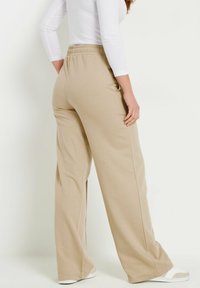 Pantalones de pierna ancha en color beige con cinturilla elástica, hechos de una tela suave. Presentan una textura lisa y bolsillos laterales, combinados con zapatillas blancas.