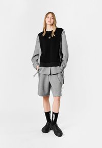 Sudadera oversized negra y gris con mangas largas, pantalones cortos grises, calzado negro texturizado y calcetines negros con letras blancas.