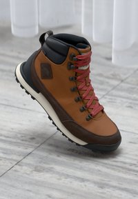 Brązowy skórzany i tekstylny but trekkingowy z czerwonymi sznurówkami, czarnym kołnierzem, trwałą gumową podeszwą i łatką z logo z boku.