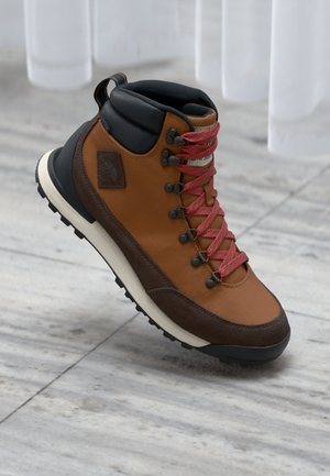 Botte de randonnée en cuir brun et textile avec des lacets rouges, un col noir, une semelle en caoutchouc durable et un patch logo sur le côté.