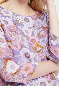 Kukallinen mekko purppurataustalla, jossa on suuria vaaleanpunaisia, valkoisia ja oransseja kukkia. Pehmeä kangas, jossa on kolme neljännestä hihoja ja pyöreä kaula-aukko.