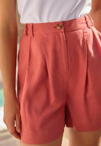 Shorts en lin rose vif avec une taille haute, un devant plissé et une fermeture à un bouton à la taille. Texture lisse et coupe ajustée.