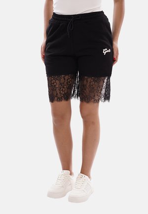 Mujer con pantalones cortos negros con ribete de encaje floral que se extiende debajo del dobladillo, zapatillas blancas y una camiseta blanca, de pie contra un fondo liso.
