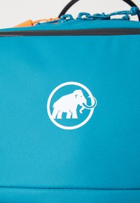 Tyrkis stoffpose med et fremtredende hvitt sirkulært logo av en mammut, med teksturert overflate og kontrastsømmer.