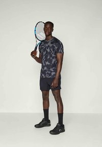 Musta kamouflage urheilupaita ja mustat shortsit; pitää kädessään tennismailaa, jonka sininen korostus. Mustat urheilukengät, joissa on teksturoitu pohja.