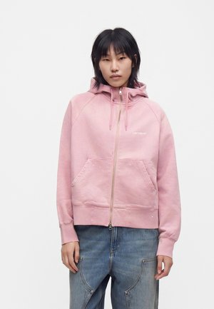 Person iført en lyserød lynlåshoodie med forlommer og lyseblå jeans, stående mod en ensfarvet hvid baggrund.