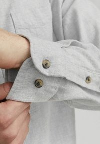 Grijze blouse met een knoopmanchet met twee bruine knopen. Getextureerde stof met stikseldetails en een borstzak.