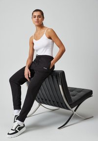 Zwarte sportbroek met een elastische tailleband, witte tanktop en zwart-witte sneakers. Het model zit op een moderne zwart-chroom stoel.