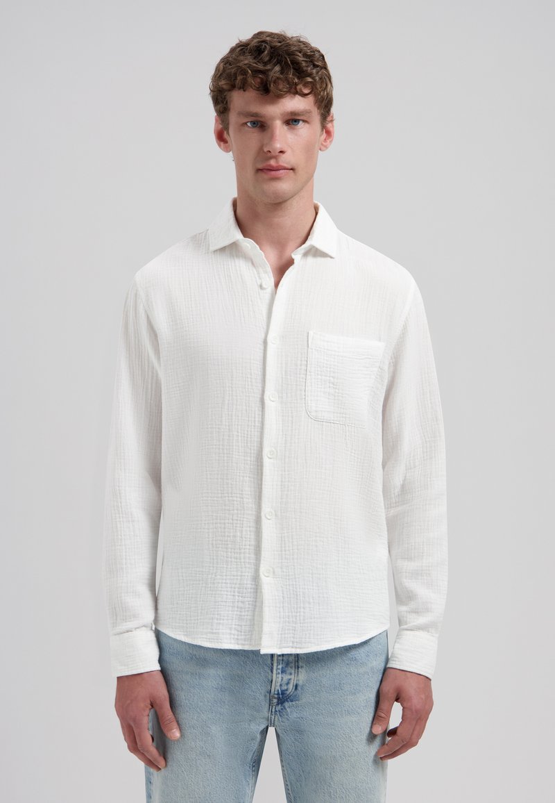 Jeune homme aux cheveux bouclés portant une chemise blanche texturée à boutons avec une poche poitrine et un jean bleu clair sur un fond uni.