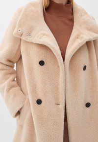 Beige dubbelknäppt kappa tillverkad av mjukt texturerat tyg, med bred krage och svarta knappar. Bärs över en brun polotröja.