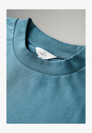 Sudadera turquesa con cuello redondo acanalado, textura suave y una etiqueta que dice "THE SET". No hay patrones ni detalles adicionales visibles.