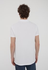 Polo shirt bianca a maniche corte, tessuto testurizzato, colletto a coste e orlo dritto, presentata su uno sfondo semplice.