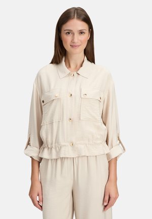 Beige kurze Jacke aus weichem Stoff, mit zwei Vordertaschen, Knopfverschluss und aufgerollten Ärmeln mit Riegel, die eine entspannte Passform schaffen.