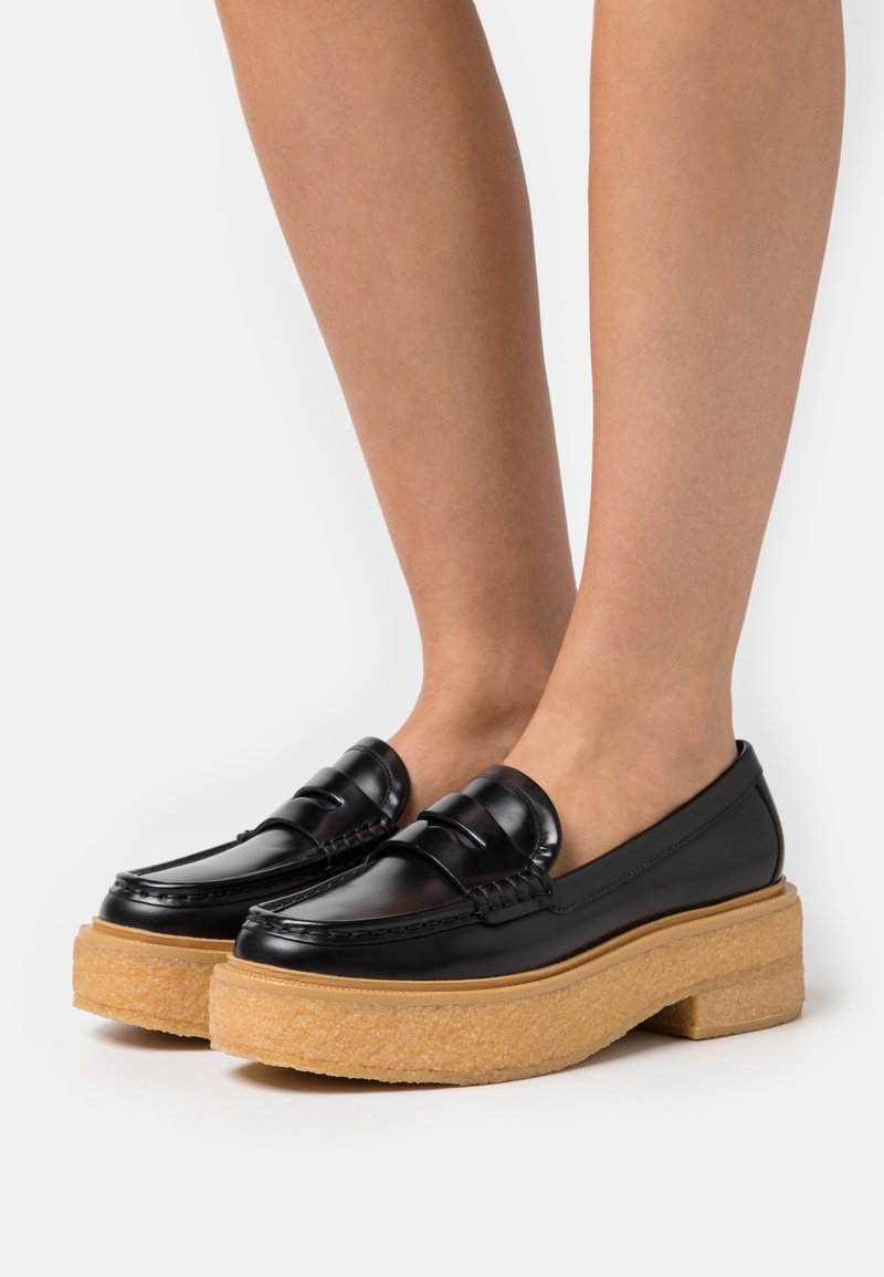 Jambes portant des mocassins à plateforme en cuir noir avec des semelles épaisses et texturées couleur sable sur fond blanc.
