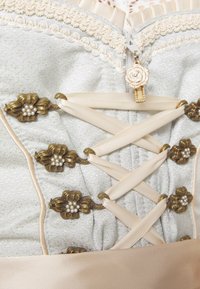 Corset à lacets crème avec fermoirs floraux en bronze, accents de perles, délicate bordure en dentelle et petit détail de tirette en forme de rose.