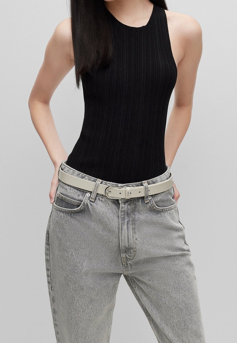 Femme portant un top noir sans manches côtelé, un jean gris taille haute et une ceinture beige clair, mains dans les poches arrière.
