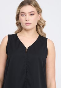 Blusa nera senza maniche con scollatura a V e un piccolo dettaglio in metallo dorato al centro. Tessuto morbido, vestibilità comoda e drappeggio sottile.