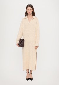 Beige adījuma kleita ar V-veida kaklu un sānu šķēlumiem. Modelis tur tumši brūnu maku un valkā melnus šaurās purngalu zābakus.