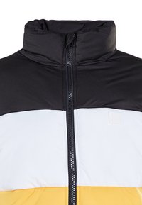Puffjakke med svart overdel, hvit midtdel og gul underdel. Høy krage, hel glidelås og glatt tekstur.