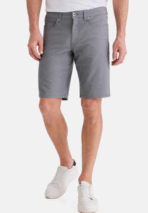 Mann trägt knielange graue Shorts und weiße Turnschuhe, steht vor einem schlichten Hintergrund.