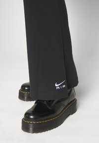 Fekete pamutnadrág fehér "AIR" logóval az alján, fényes fekete platformcsizmával párosítva, sárga varrással és texturált talppal.