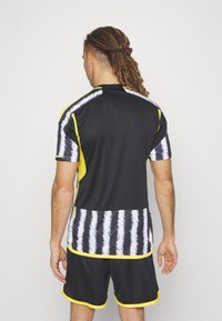 Maglia atletica nera con accenti gialli e motivo a strisce bianche sul retro. Il materiale sembra liscio e leggero. Abbinata a pantaloni corti neri.