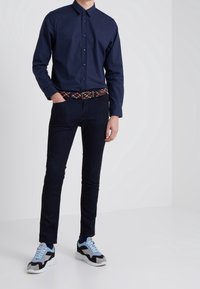 Námořnická košile s knoflíky, tmavé džíny, vzorovaný opasek a vícebarevné tenisky s akcenty, které zahrnují kombinaci materiálů a textur.