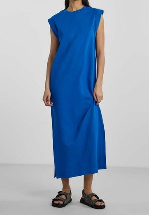 Nainen, joka pukeutunut hihattomaan siniseen midimekkoon, jossa on toppatut olkapäät, ja mustiin avoimiin varvassandaaleihin, yksinkertaisella harmaalla taustalla.