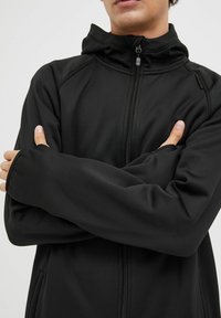 Crna jakna na zip s visokim ovratnikom i prilagođenim manžetama, napravljena od glatkog materijala s teksturiranim završetkom i minimalnim detaljima od hardvera.