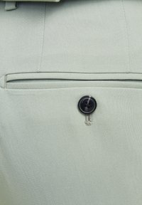 Lysegrønn stoff med teksturert finish, med en svart knapp og en sydd lomme detalj. Glatte, skreddersydde kanter synlige.