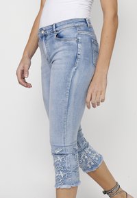 Light blue denim capri jeans featuring frayed hems and white floral embroidery at the cuffs, with a slim-fit design and classic five-pocket style.