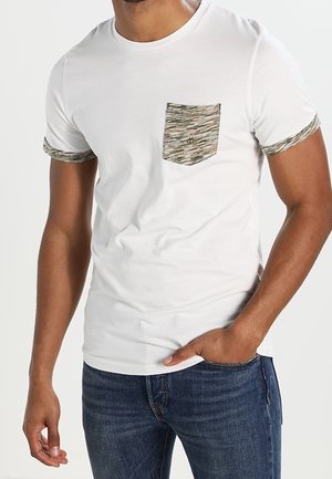 Mężczyzna w białym t-shirt z krótkim rękawem z brązową kieszenią i mankietami z wzorem, w połączeniu z niebieskimi dżinsami.