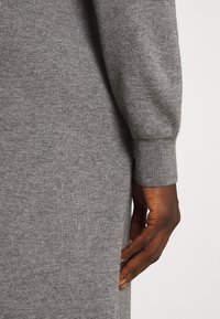 Main droite d'une personne à la peau foncée reposant à côté d'un vêtement en tricot gris à manches longues avec des poignets côtelés, sur un fond neutre.