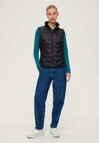 Schwarze gesteppte Weste mit hohem Kragen, getragen über einem türkisfarbenen Langarmshirt. Kombiniert mit blauen Jeans und weißen Sneakers. Einfaches Design.