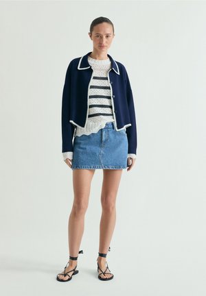 Donna che indossa un cardigan corto blu navy con bottoni, maglione bianco a righe, minigonna di jeans e sandali neri con cinturini, in piedi su sfondo semplice.