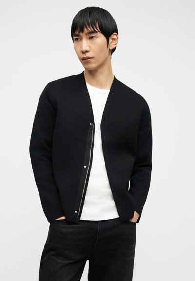 Άνδρας με μαύρη ζακέτα με φερμουάρ πάνω από λευκό πουκάμισο και μαύρα τζιν, όρθιος με τα χέρια στις τσέπες μπροστά σε απλό φόντο.