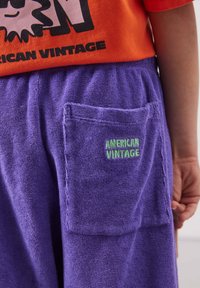 Enfant portant un t-shirt orange et un short en éponge violet avec « American Vintage » brodé sur la poche arrière, main reposant sur le côté.