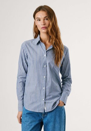 Camicia a righe blu e bianche con maniche lunghe, colletto classico e dettaglio della tasca frontale. Indossata con jeans blu a vestibilità comoda.
