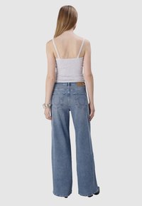 Lichtgrijze tanktop met dunne schouderbandjes, gecombineerd met blauwe jeans met wijde pijpen. De jeans hebben achterzakken en een bruine merklabel op de taille.