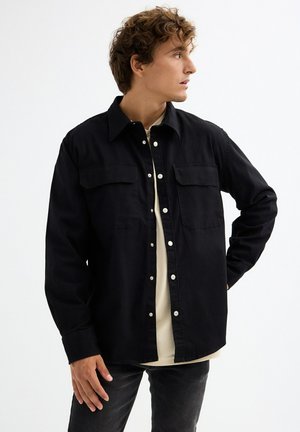 Camisa de algodón negra con cuello puntiagudo, dos bolsillos en el pecho, botones plateados y mangas largas, llevada sobre una camisa de color claro.