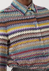 Närbild av en knäppskjorta med flerfärgad zickzack-mönstring, metalltrådar och spetsig krage.