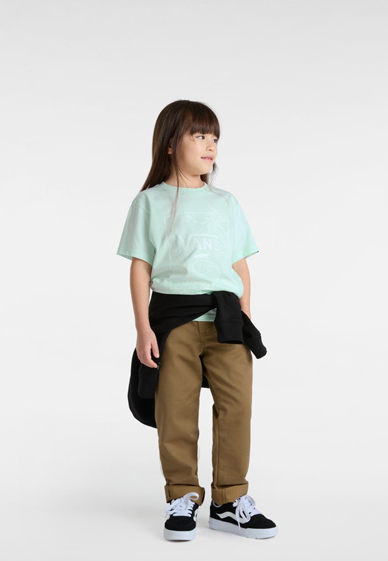 Minzgrünes T-Shirt mit kurzen Ärmeln und weißem Grafiklogo, kombiniert mit braunen Hosen und schwarz-weißen Turnschuhen. Schwarzes Sweatshirt in der Taille gebunden.
