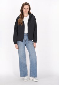 Giacca nera leggera con cappuccio, polsini elastici e tasche frontali, abbinata a jeans blu a vita alta e gamba larga, insieme a sneakers bianche.