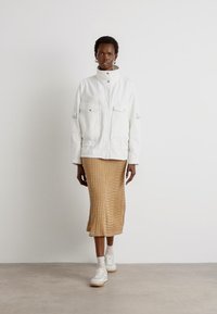White, zip-up jacket with two front pockets, worn over a textured, ribbed, beige midi skirt. White sneakers complete the outfit.