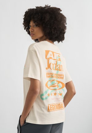 Kobieta o kręconych włosach, ubrana w kremowy t-shirt z grafiką Air Jordan World Tour i ciemne spodnie, pozująca na jasnym, jednolitym tle.