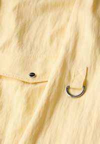 Tessuto giallo con una superficie strutturata e a coste. Presenta un anello in metallo argentato e un bottone a pressione, creando un elemento di design funzionale.