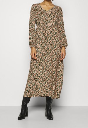 Femme portant une robe midi fleurie à manches longues avec un décolleté en V et des bottes noires à talons, debout devant un fond neutre.