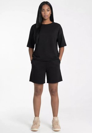 Kobieta z długimi czarnymi włosami, ubrana w czarną koszulkę z krótkim rękawem i czarne szorty, beżowe trampki, stojąca z rękami w kieszeniach na białym tle.