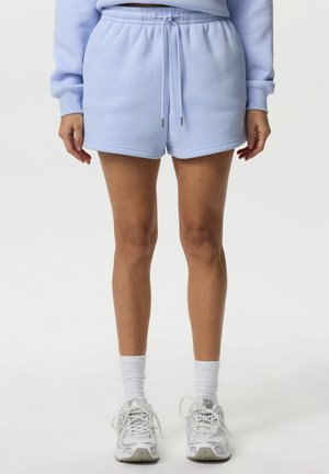 Osoba nosząca jasnoniebieskie elastyczne szorty ze ściągaczem w talii, białe skarpetki sportowe i białe sportowe buty, stojąca na białym tle.